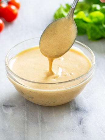 A spoon scooping out Big Mac Sauce from a small glass bowl with lettuce and tomatoes in the background.