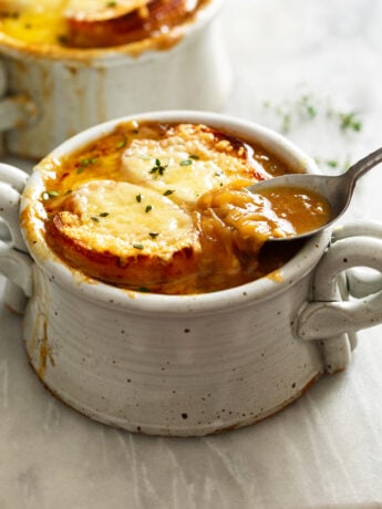 French Onion Soup in a crock with melted cheese and bread on top and a spoon scooping up the broth.